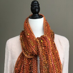 K&ouml;nnte beinhalten: Ein orange-brauner Strickschal mit gelben und rosa Akzenten. Der Schal ist &uuml;ber einer Schaufensterpuppe drapiert, was seine Textur und Farbvariationen hervorhebt. Der handgefertigte Schal wirkt warm und gem&uuml;tlich.