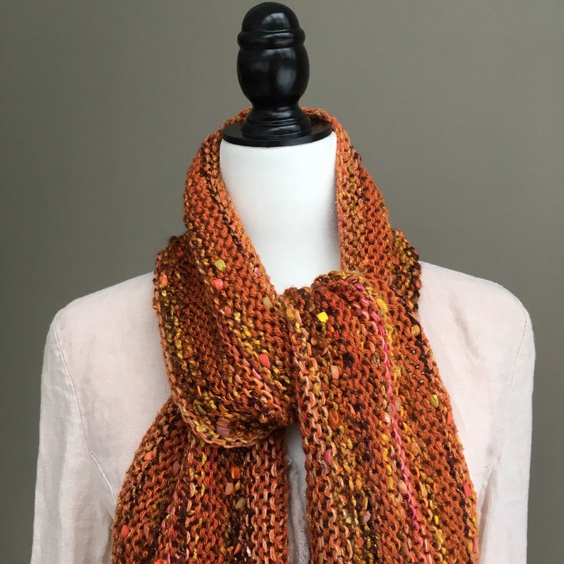 K&ouml;nnte beinhalten: Ein orange-brauner Strickschal mit gelben und rosa Akzenten. Der Schal ist &uuml;ber einer Schaufensterpuppe drapiert, was seine Textur und Farbvariationen hervorhebt. Der handgefertigte Schal wirkt warm und gem&uuml;tlich.