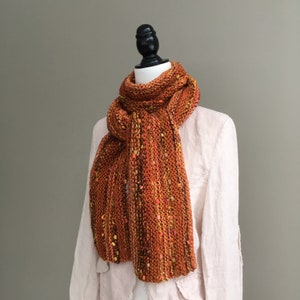 K&ouml;nnte beinhalten: Handgestrickter Schal in Orange-, Braun- und Gelbt&ouml;nen, pr&auml;sentiert auf einer Schaufensterpuppe. Der Schal hat eine grob gestrickte Textur mit Farbtupfern. Er ist drapiert, um seine L&auml;nge und sein Design zu zeigen. Der Schal ist ca. 180 cm lang.