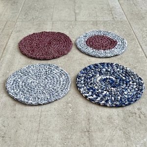 Puede incluir: Cuatro posavasos circulares hechos a mano en tonos burdeos, grises y azules. Los posavasos están hechos de un material texturizado y tejido apretado, con un patrón en espiral. Los posavasos tienen aproximadamente 10 cm de diámetro.