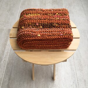 K&ouml;nnte beinhalten: Ein rechteckiger, handgestrickter Gegenstand in Braun-, Orange- und Gelbt&ouml;nen mit Farbtupfern. Er liegt auf einem kleinen, runden, hellen Holztisch vor einem grau gefliesten Boden. Das Objekt scheint ein Kissen zu sein.