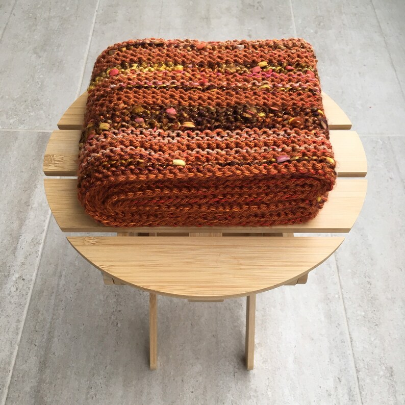 K&ouml;nnte beinhalten: Ein rechteckiger, handgestrickter Gegenstand in Braun-, Orange- und Gelbt&ouml;nen mit Farbtupfern. Er liegt auf einem kleinen, runden, hellen Holztisch vor einem grau gefliesten Boden. Das Objekt scheint ein Kissen zu sein.