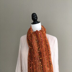 K&ouml;nnte beinhalten: Ein orange-brauner Strickschal mit gelben und rosa Flecken. Der Schal ist auf einer Schaufensterpuppe drapiert, um seine Textur und Farbvariationen zu zeigen. Das Design ist handgestrickt und hat eine grobe Struktur.