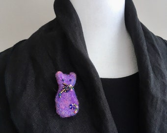 May include: Purple felt cat brooch with beaded details. The cat is sitting upright with its tail curled around its body.