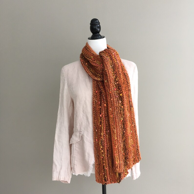 K&ouml;nnte beinhalten: Ein gestrickter Schal in Orange- und Braunt&ouml;nen, gesprenkelt mit Gelb und Rosa, wird auf einer Schaufensterpuppe pr&auml;sentiert. Der Schal aus dickem Garn hat eine strukturierte Optik. Er ist &uuml;ber einer hellrosa Leinenjacke drapiert.