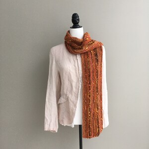 K&ouml;nnte beinhalten: Ein orange-gelber Strickschal, der auf einer Schaufensterpuppe drapiert ist. Der Schal hat eine strukturierte Optik mit gelben und orangenen Flecken. Die Schaufensterpuppe tr&auml;gt einen hellfarbigen Blazer.
