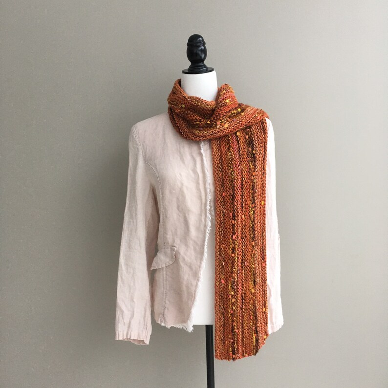 K&ouml;nnte beinhalten: Ein orange-gelber Strickschal, der auf einer Schaufensterpuppe drapiert ist. Der Schal hat eine strukturierte Optik mit gelben und orangenen Flecken. Die Schaufensterpuppe tr&auml;gt einen hellfarbigen Blazer.