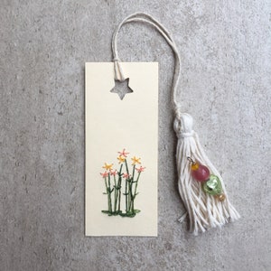 以下が含まれることがあります： 星形の切り抜きとピンク、イエロー、グリーンの刺繍の花が施されたベージュのしおり。しおりには、ピンクとグリーンのビーズが付いた白いタッセルが付いています。