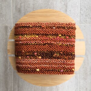 K&ouml;nnte beinhalten: Ein handgestricktes, strukturiertes Stirnband in Braun-, Orange- und Rosat&ouml;nen mit gelben und anderen farbigen Flecken. Das Stirnband liegt auf einem runden, hellen Holzhalter.