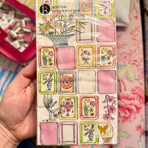 K&ouml;nnte beinhalten: Eine Packung dekorativer Papierservietten mit floralem und botanischem Design. Die Servietten haben ein Rastermuster mit rosa Quadraten und Aquarellillustrationen von Blumen, Vasen und V&ouml;geln. Der Text "BOSTON INTERNATIONAL" ist sichtbar.