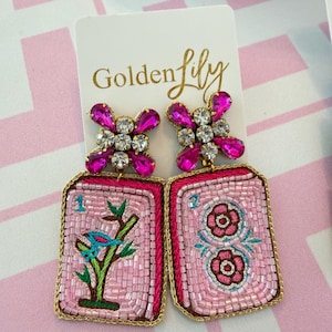 May include: A pair of rectangular beaded earrings on a Golden Lily display card. Each earring features a pink beaded base with gold trim, topped with a cluster of clear and fuchsia crystals. One earring shows a bird on a branch, the other two flowers.