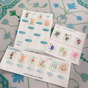 May include: Three sets of clear acrylic drink accessories in plastic packaging. One set contains four 6.25-inch stir sticks, one set contains six 3-inch party toppers, and one set contains two drink clips, all featuring colorful mahjong tile designs.