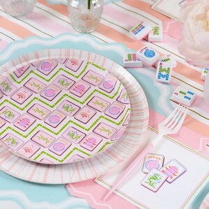 May include: A decorative table setting featuring a pink and white striped tablecloth, a scalloped edge plate, and a matching plate with a pink and green Mahjong tile design. Mahjong tiles and a white fork are scattered around.
