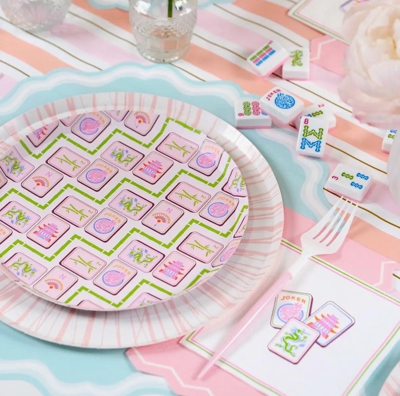 May include: A decorative table setting featuring a pink and white striped tablecloth, a scalloped edge plate, and a matching plate with a pink and green Mahjong tile design. Mahjong tiles and a white fork are scattered around.