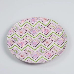 May include: A round, decorative plate with a repeating pattern of pink and green geometric shapes. The pattern includes images of dragons, fans, pagodas, and the word "Joker". The plate has a white background.