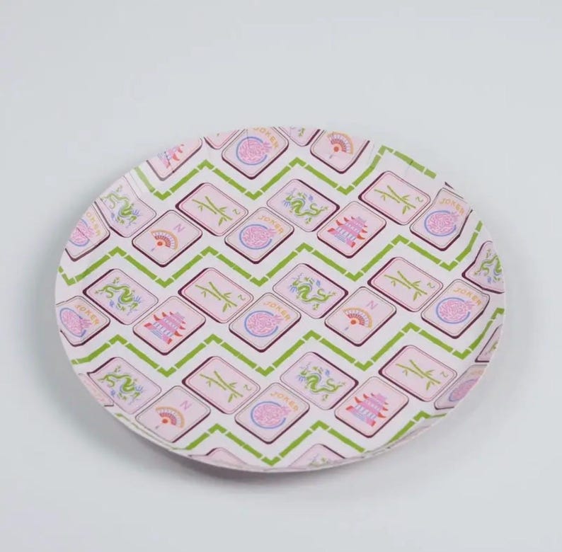 May include: A round, decorative plate with a repeating pattern of pink and green geometric shapes. The pattern includes images of dragons, fans, pagodas, and the word "Joker". The plate has a white background.