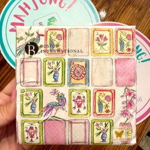 K&ouml;nnte beinhalten: Ein quadratisches Papierservietten-Set mit einem Aquarelldesign mit gerahmten Bildern von Blumen, Vasen und Laternen. Das Servietten-Set ist mit "Boston International" beschriftet. Zwei Untersetzer mit dem Wort "Mahjong" befinden sich im Hintergrund.