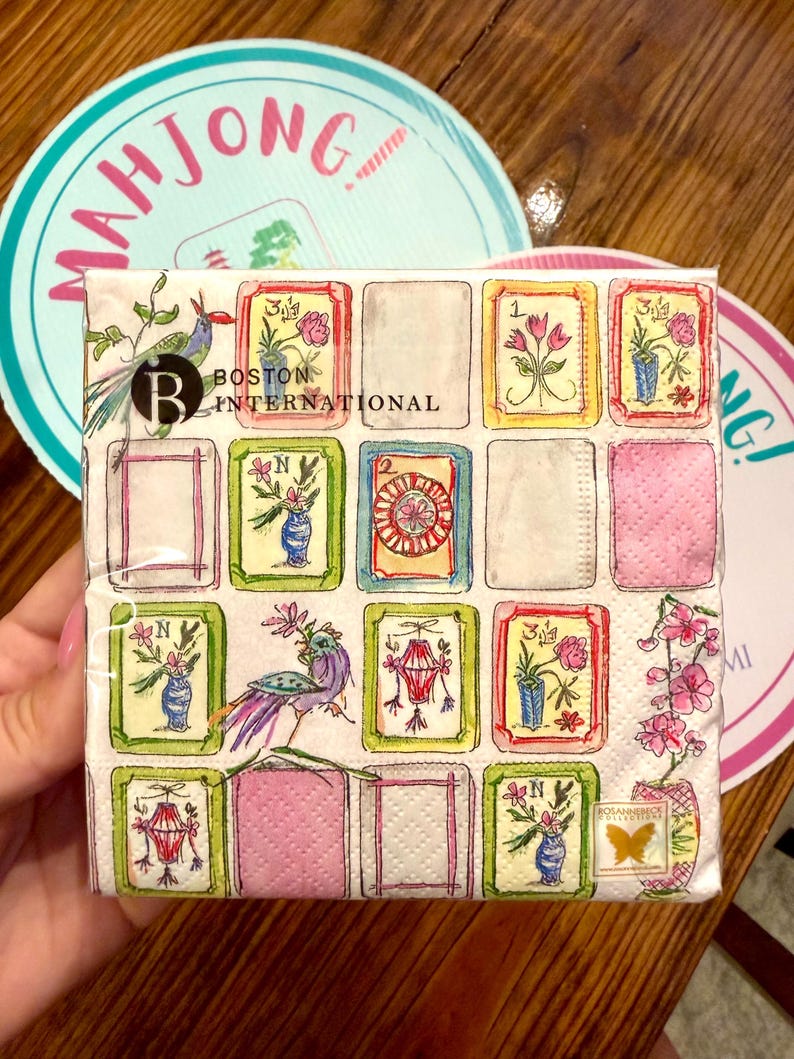 K&ouml;nnte beinhalten: Ein quadratisches Papierservietten-Set mit einem Aquarelldesign mit gerahmten Bildern von Blumen, Vasen und Laternen. Das Servietten-Set ist mit "Boston International" beschriftet. Zwei Untersetzer mit dem Wort "Mahjong" befinden sich im Hintergrund.