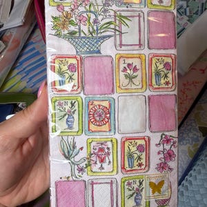 K&ouml;nnte beinhalten: Ein Paket dekorativer Papierservietten mit floralem Design. Die Servietten zeigen ein Raster aus farbigen Quadraten mit floralen Illustrationen, rosa Rechtecken und dem Text "BOSTON INTERNATIONAL". Das Logo der Rosannebeck Collection ist ebenfalls sichtbar.