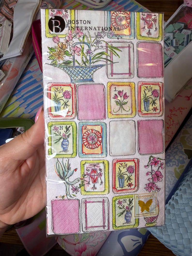 K&ouml;nnte beinhalten: Ein Paket dekorativer Papierservietten mit floralem Design. Die Servietten zeigen ein Raster aus farbigen Quadraten mit floralen Illustrationen, rosa Rechtecken und dem Text "BOSTON INTERNATIONAL". Das Logo der Rosannebeck Collection ist ebenfalls sichtbar.