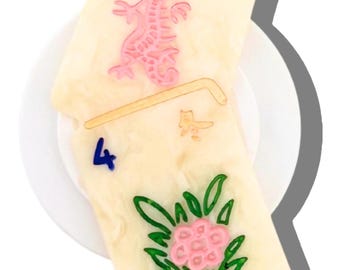 May include: Two rectangular off-white tiles with engraved designs. The top tile features a pink dragon, while the bottom tile displays a blue number 4, a small gold character, and a pink flower with green leaves, all resting on a white plate.
