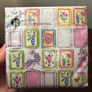 K&ouml;nnte beinhalten: Eine Packung dekorativer Papierservietten mit einem sich wiederholenden Muster aus gerahmten Blumen- und Laternenillustrationen. Die Servietten haben einen wei&szlig;en Hintergrund mit rosa und gr&uuml;nen Akzenten, und der Text "BOSTON INTERNATIONAL" ist sichtbar.