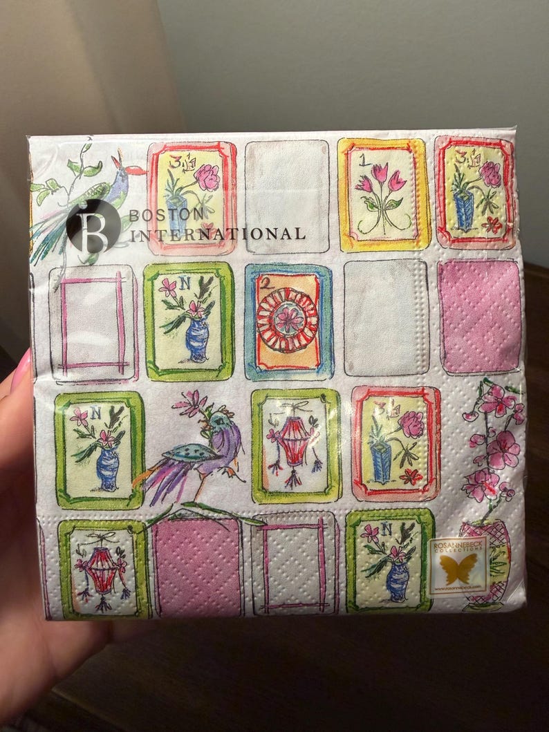 K&ouml;nnte beinhalten: Eine Packung dekorativer Papierservietten mit einem sich wiederholenden Muster aus gerahmten Blumen- und Laternenillustrationen. Die Servietten haben einen wei&szlig;en Hintergrund mit rosa und gr&uuml;nen Akzenten, und der Text "BOSTON INTERNATIONAL" ist sichtbar.