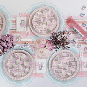 May include: A festive table setting with pink and green patterned paper plates, napkins, and cutlery. The plates and napkins feature a repeating design. Scattered mahjong tiles and flowers add to the celebratory atmosphere. The table is decorated with pink and blue scalloped edges.