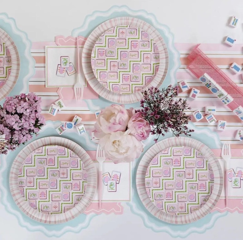 May include: A festive table setting with pink and green patterned paper plates, napkins, and cutlery. The plates and napkins feature a repeating design. Scattered mahjong tiles and flowers add to the celebratory atmosphere. The table is decorated with pink and blue scalloped edges.