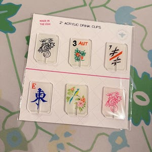 May include: A set of six clear 2-inch acrylic drink clips on a white display card. The clips feature various designs including a black dragon, the text 3 AUT, a black and orange symbol, the letter E with a blue character, a bird with flowers, and a pink dragon.