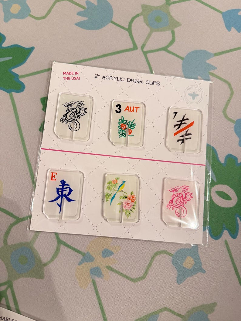 May include: A set of six clear 2-inch acrylic drink clips on a white display card. The clips feature various designs including a black dragon, the text 3 AUT, a black and orange symbol, the letter E with a blue character, a bird with flowers, and a pink dragon.