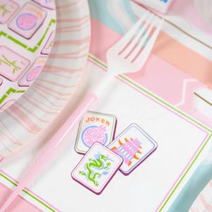 K&ouml;nnte beinhalten: Eine rosa und wei&szlig;e Party-Szene mit Mahjong-Thema. Das Bild zeigt eine wei&szlig;e Plastikgabel, Pappteller und eine Serviette mit drei Mahjong-Steinen. Die Steine zeigen einen Drachen, eine Pagode und das Wort "JOKER".
