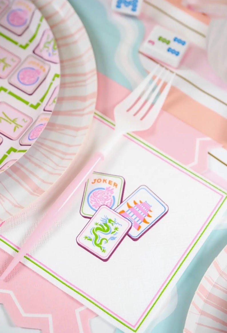 K&ouml;nnte beinhalten: Eine rosa und wei&szlig;e Party-Szene mit Mahjong-Thema. Das Bild zeigt eine wei&szlig;e Plastikgabel, Pappteller und eine Serviette mit drei Mahjong-Steinen. Die Steine zeigen einen Drachen, eine Pagode und das Wort "JOKER".