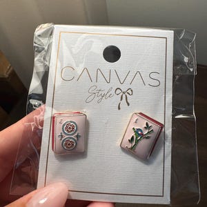May include: A pair of pink enamel earrings with red trim, one featuring a floral design and the other a bird on a branch. The earrings are displayed on a white card with the text "CANVAS Style".
