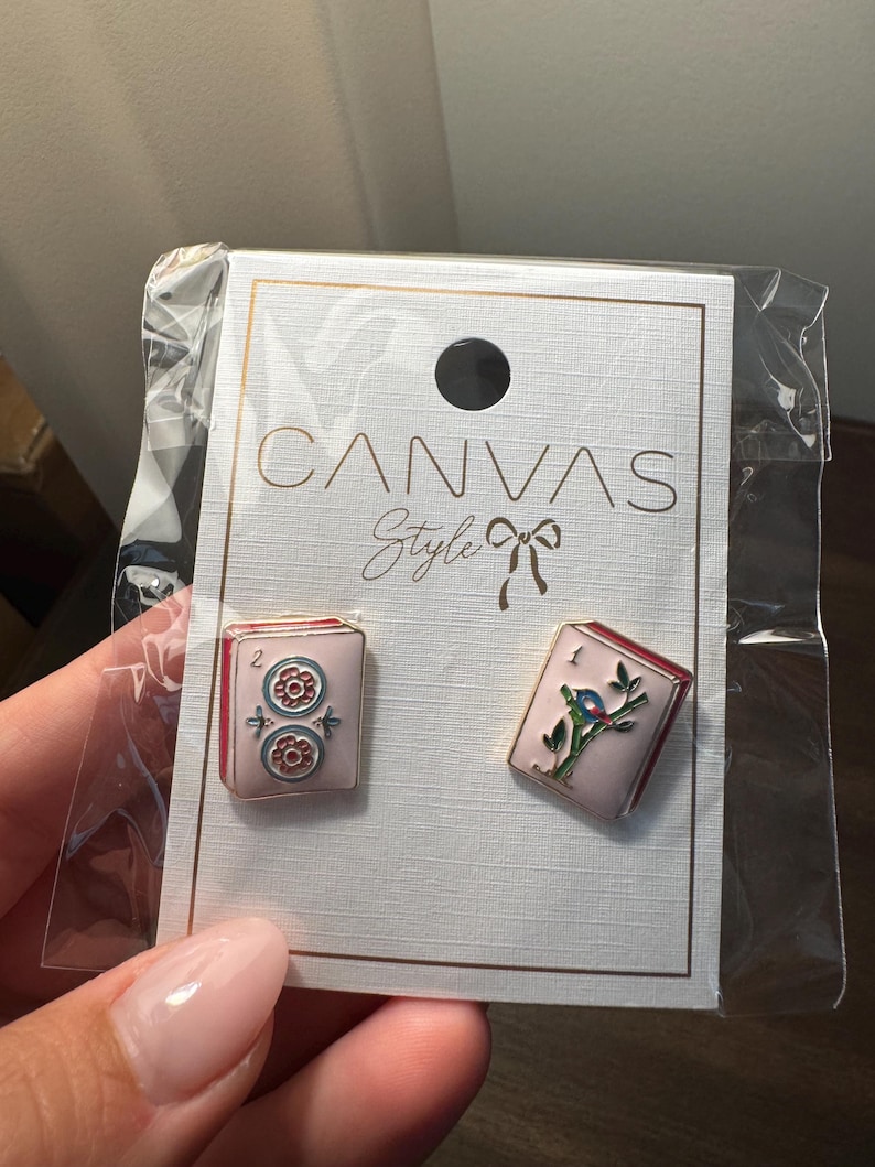 May include: A pair of pink enamel earrings with red trim, one featuring a floral design and the other a bird on a branch. The earrings are displayed on a white card with the text "CANVAS Style".