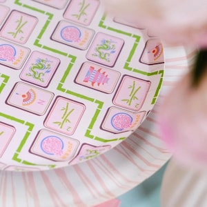 May include: A close-up of a pink and white paper plate with a Mahjong tile pattern. The tiles are pink with green and white accents. The plate is on top of a striped pink and white plate.