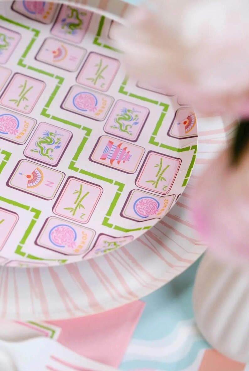 May include: A close-up of a pink and white paper plate with a Mahjong tile pattern. The tiles are pink with green and white accents. The plate is on top of a striped pink and white plate.
