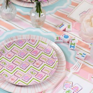 K&ouml;nnte beinhalten: Eine Tischdekoration mit rosa und blau gestreiften Tischsets, Tellern und Mahjong-Steinen. Die Teller haben ein rosa-gr&uuml;nes Kachelmuster. Kleine Vasen halten rosa Blumen. Eine wei&szlig;e Gabel und dekorative Karten sind ebenfalls vorhanden.