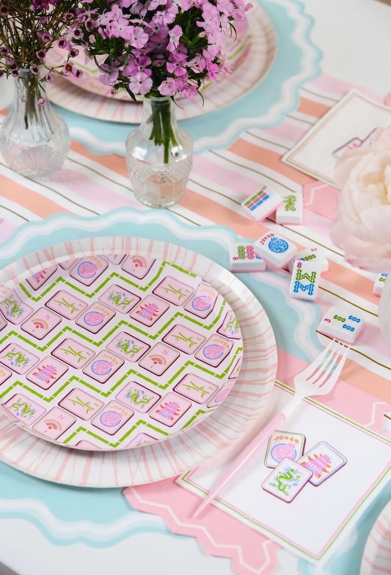 K&ouml;nnte beinhalten: Eine Tischdekoration mit rosa und blau gestreiften Tischsets, Tellern und Mahjong-Steinen. Die Teller haben ein rosa-gr&uuml;nes Kachelmuster. Kleine Vasen halten rosa Blumen. Eine wei&szlig;e Gabel und dekorative Karten sind ebenfalls vorhanden.