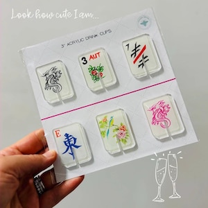 May include: A set of six clear 3-inch acrylic drink clips attached to a white display card. The clips feature various designs including a black dragon, a floral pattern with the text 3 AUT, a black and red symbol, the letter E with a blue character, a bird on a branch, and a pink dragon. The card includes the text Look how cute I am and 3 ACRYLIC DRINK CLIPS.