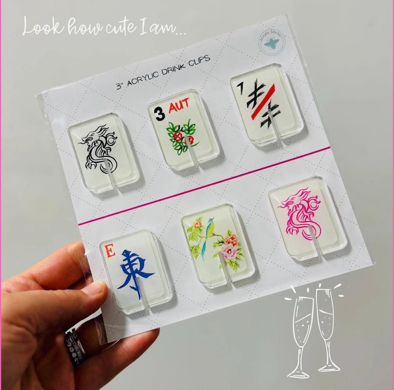 May include: A set of six clear 3-inch acrylic drink clips attached to a white display card. The clips feature various designs including a black dragon, a floral pattern with the text 3 AUT, a black and red symbol, the letter E with a blue character, a bird on a branch, and a pink dragon. The card includes the text Look how cute I am and 3 ACRYLIC DRINK CLIPS.