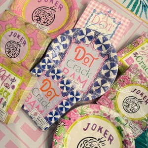 May include: A collection of paper plates and napkins featuring various designs. Some items display the text Joker, while others feature the text Dot Crack Bam. The products are packaged in clear plastic and showcase pink, blue, and green floral patterns.