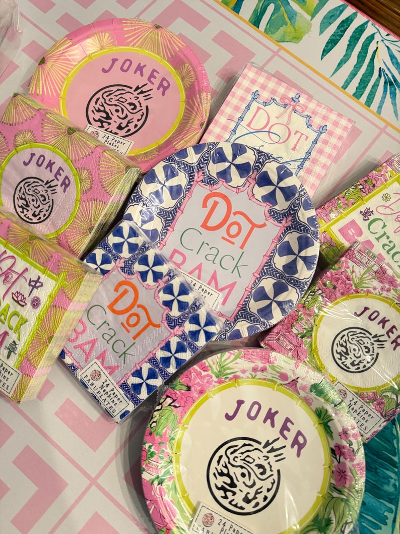 May include: A collection of paper plates and napkins featuring various designs. Some items display the text Joker, while others feature the text Dot Crack Bam. The products are packaged in clear plastic and showcase pink, blue, and green floral patterns.