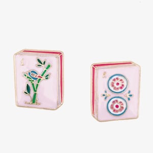 May include: Two pink Mahjong tile-shaped earrings. One earring features a bird on a bamboo branch, the other has two flower designs. Both have gold trim and red edges. The earrings are set against a white background.