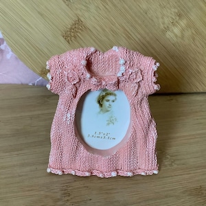 May include: A pink, dress-shaped photo frame with a textured knit design. The frame has a white border and holds a small oval portrait. The frame measures 1.5" x 2" (3.8cm x 5.1cm).