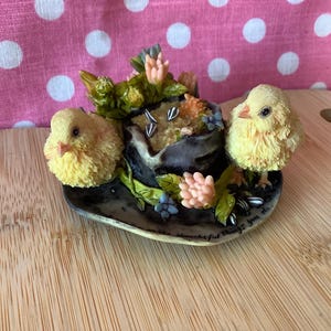 May include: A ceramic figurine of a black hat with two yellow chicks perched on the brim. The hat is decorated with flowers and greenery. The base of the figurine reads "Wonderful things come in small packages".