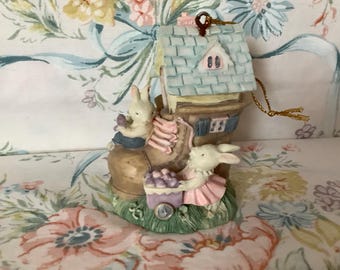 May include: A ceramic Easter ornament featuring two bunnies in a brown boot house. The house has a blue roof with a white window and a gold string for hanging.