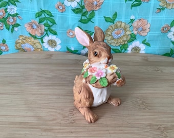 May include: A brown ceramic bunny figurine with a white belly and pink and yellow flowers around its neck. The bunny is holding a bouquet of flowers in its paws.