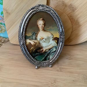 May include: An oval, silver-toned picture frame with a portrait of a woman in a white dress and blue shawl. The frame has a decorative rope-like border and rests on small feet. The background is a light wood surface.