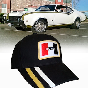 Puede incluir: Gorra de béisbol negra con una banda blanca y dorada en el borde. La gorra tiene un parche bordado rojo y negro con la palabra "HURST" en letras blancas.